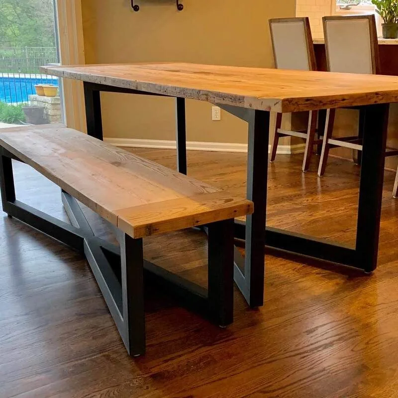 Modern wooden dining table with a thick natural wood top and black metal frame legs, paired with a matching bench, photographed against a dark studio wall.