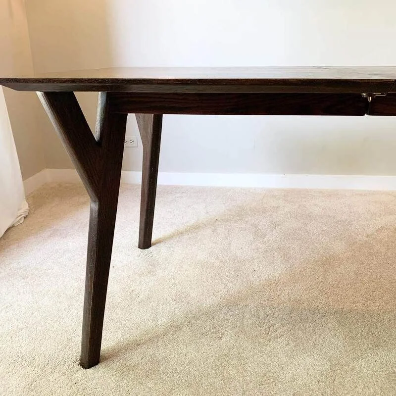 Close-up of a dark-finished wooden table with V-shaped angled legs, placed on a light carpeted floor against a white wall.