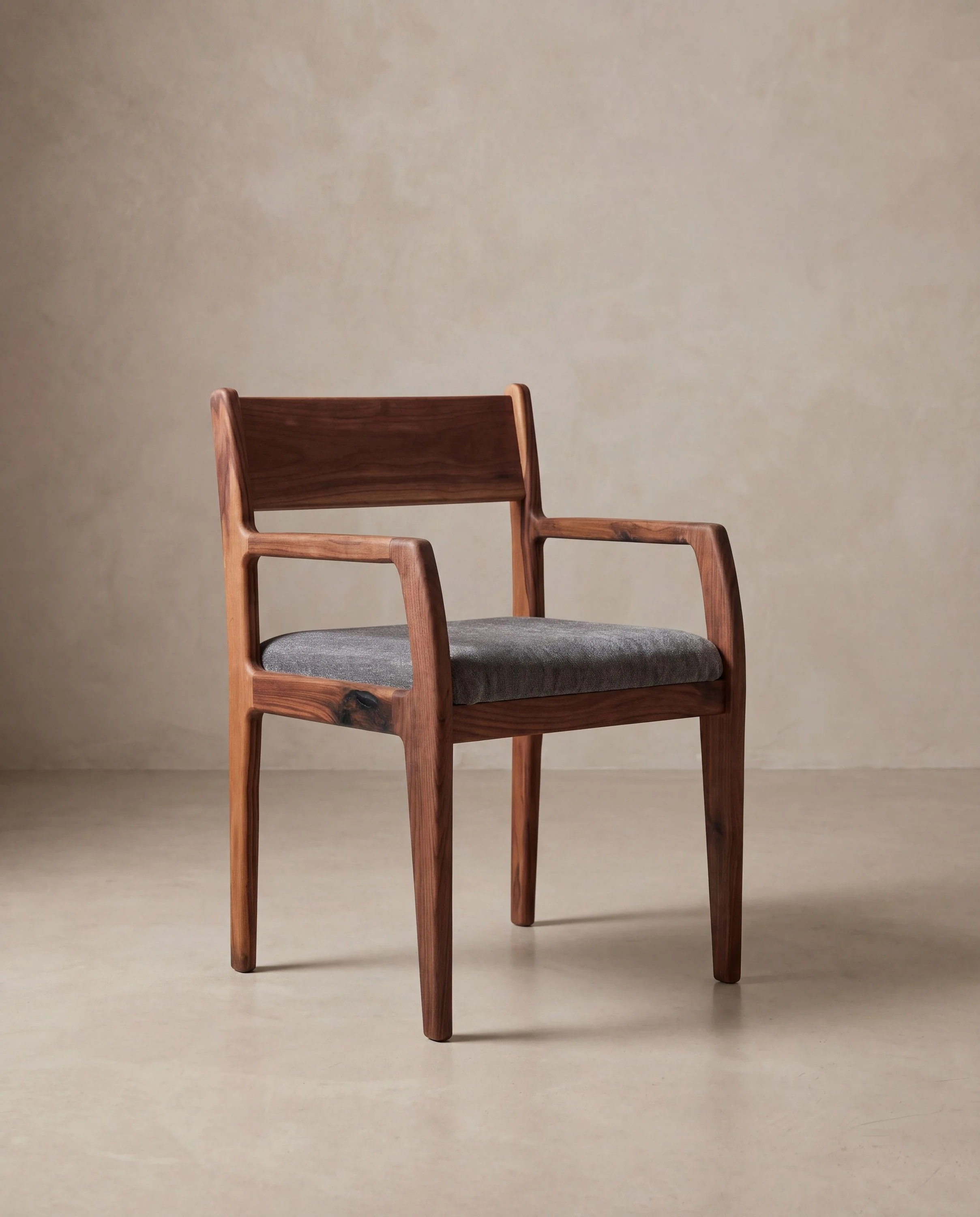 A rendering of a walnut armchair with shaped arms and a grey upholstered seat, in a limewash studio.