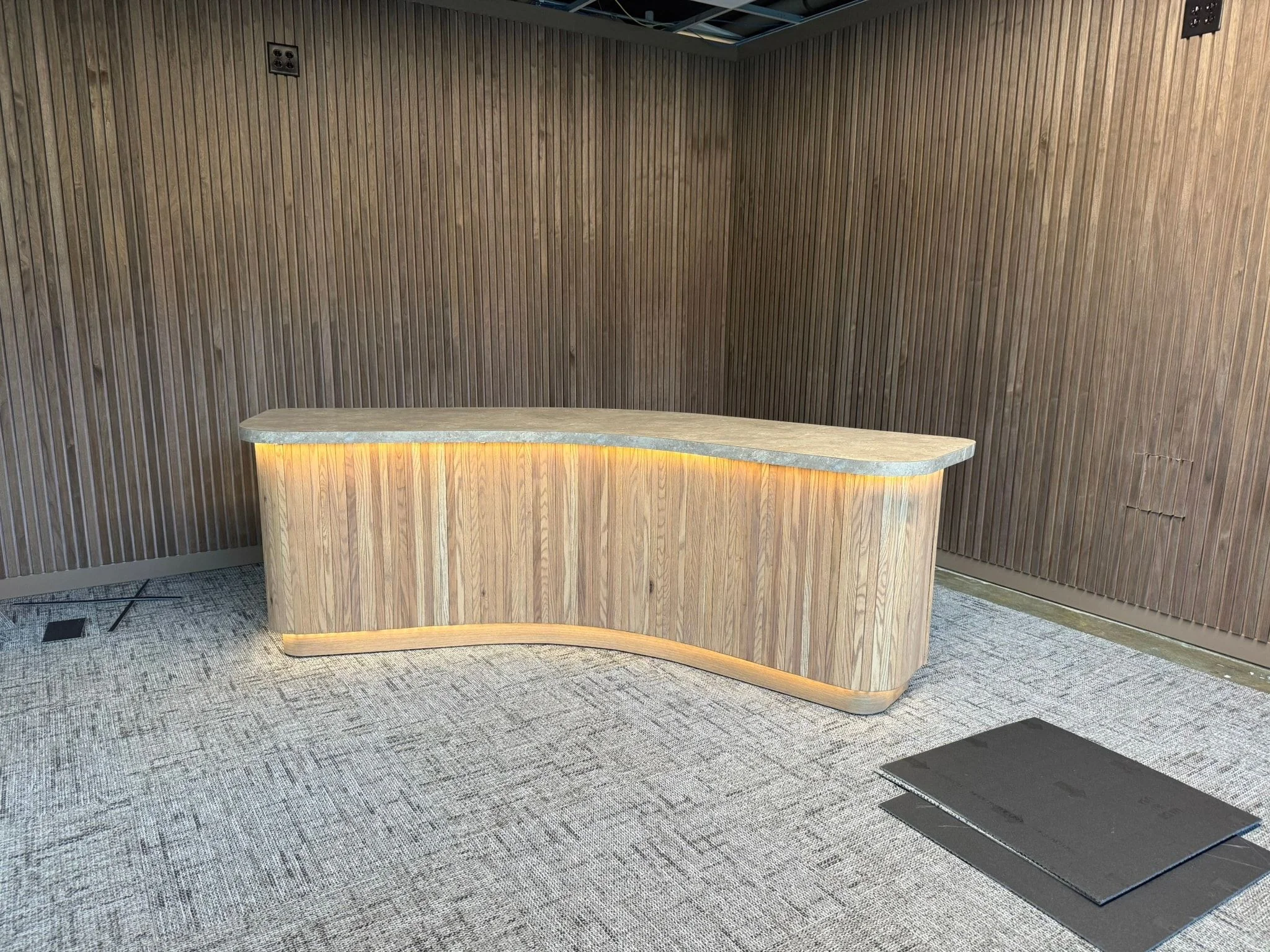 A freestanding curved desk with a slatted wood front and a light strip beneath the top, in a room lined with vertical wood slats.