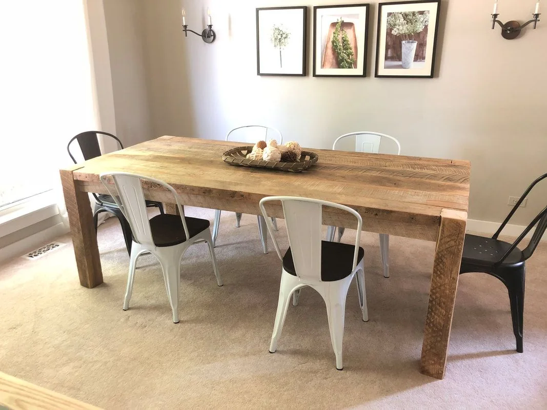 Rustic wooden dining table with six metal chairs, decorative tray with spheres, beige carpet, botanical prints, and wall sconces in a bright room.