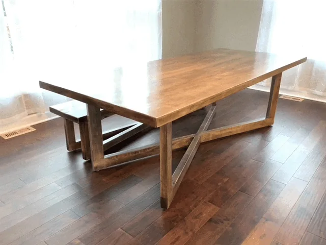 Handcrafted wooden dining table with matching bench featuring X-shaped base, photographed against a dark studio wall.