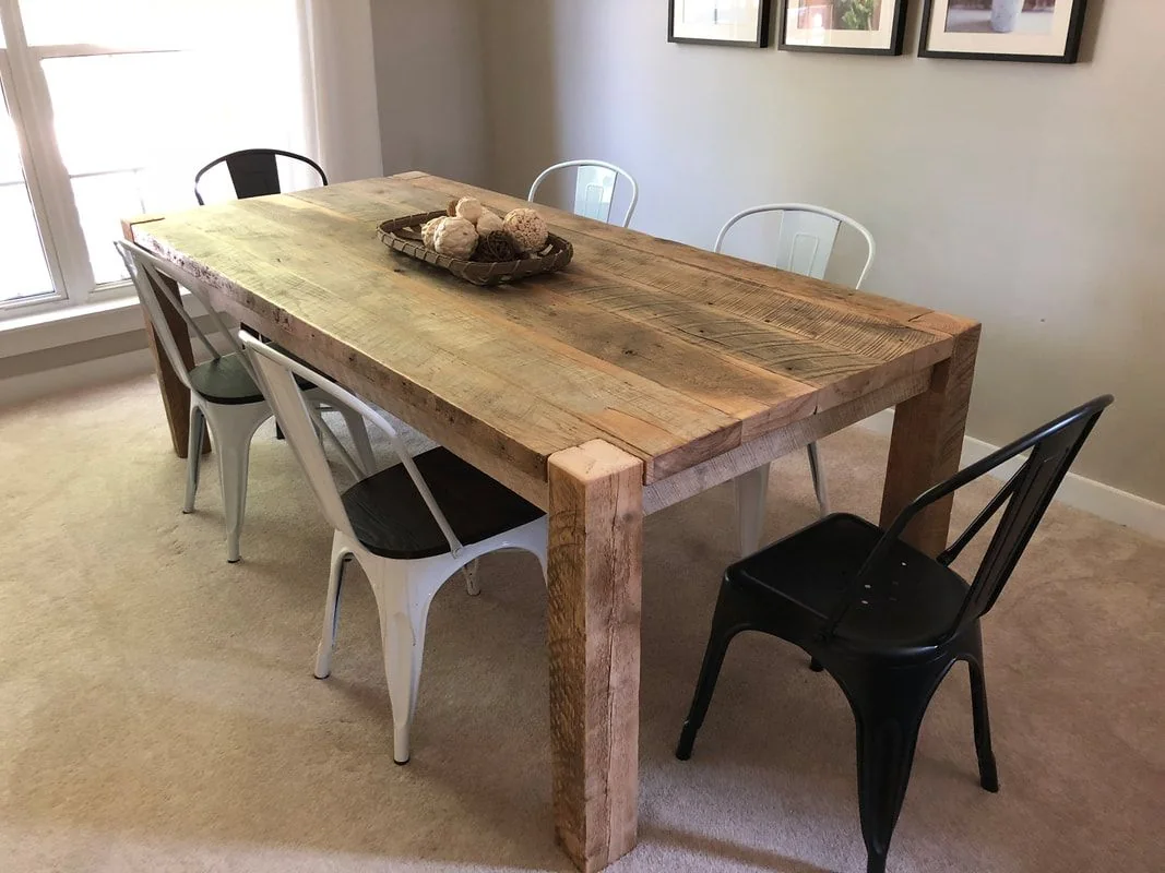 Rustic wooden dining table with six metal chairs, decorative tray with spheres, in a bright room with large windows and framed wall art.