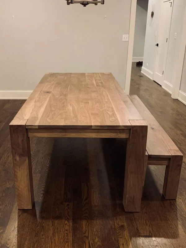 Wooden dining table with matching bench in a minimalist room with hardwood flooring and neutral walls.
