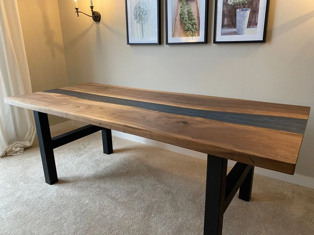 A wooden table with black legs and a dark wavy stripe across the center, photographed against a dark studio wall.