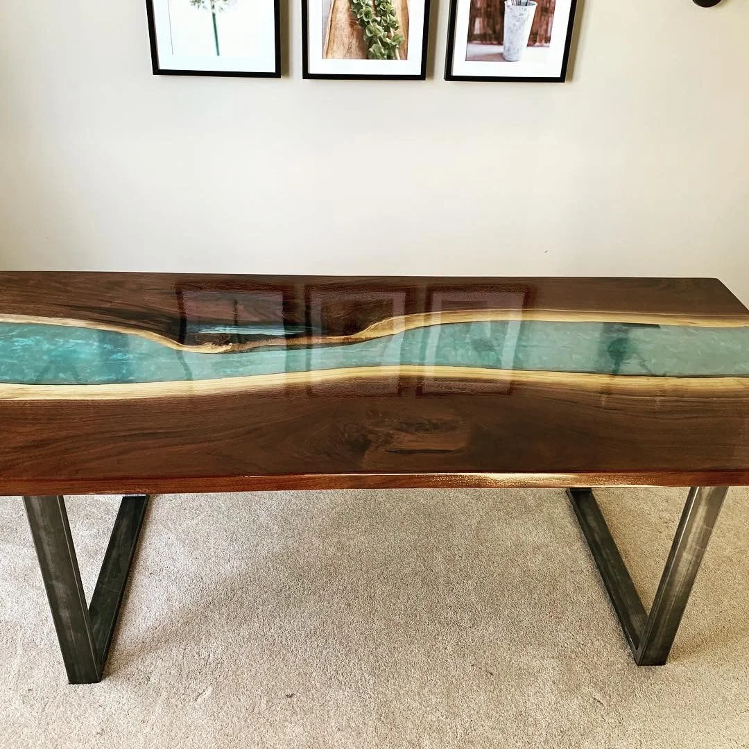 A wooden table with a blue resin river design running through the center, flanked by natural wood edges, photographed against a dark studio wall.
