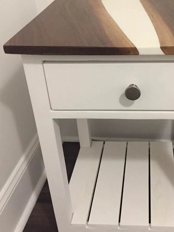 A white wooden side table with a single drawer featuring a round metallic knob, a slatted lower shelf, and a tabletop with a wave-like design combining dark wood and a light-colored inlay.