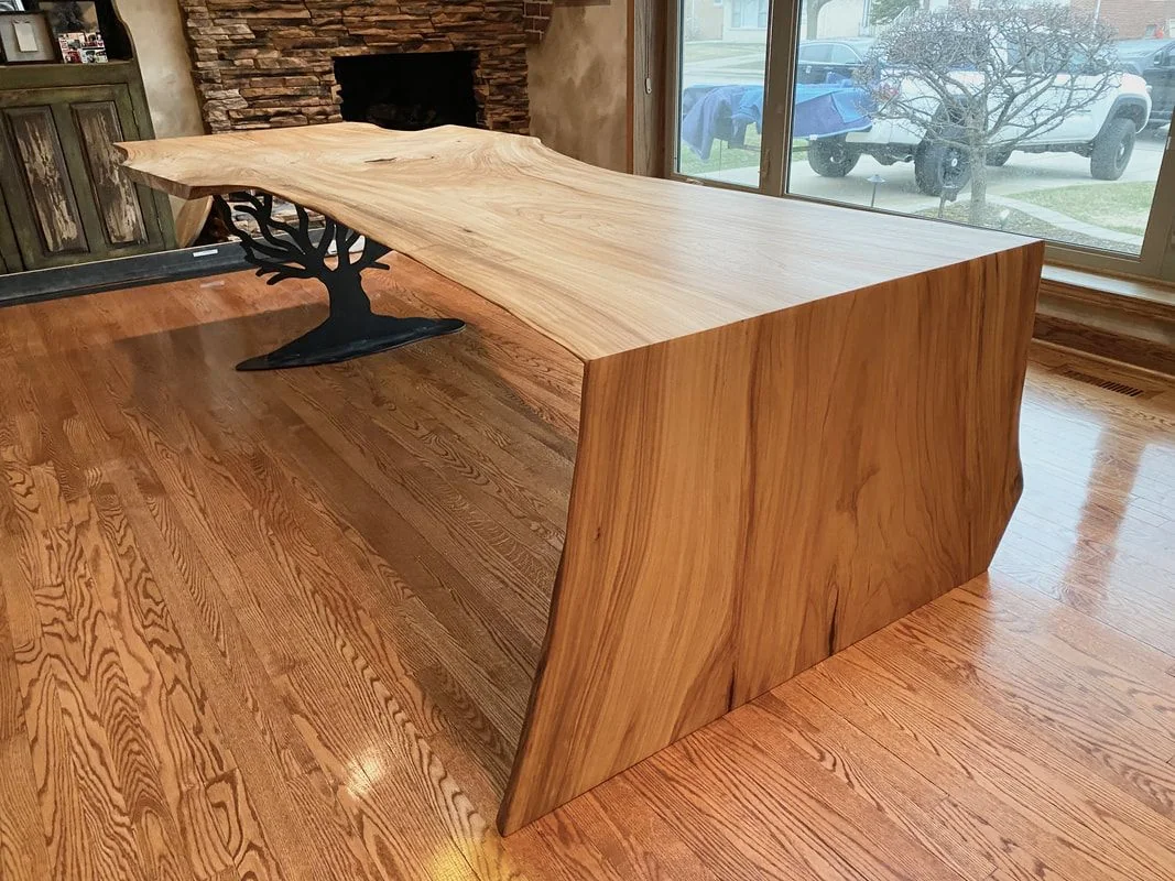 Live edge solid wood table with artistic metal tree base, photographed against a dark studio wall.