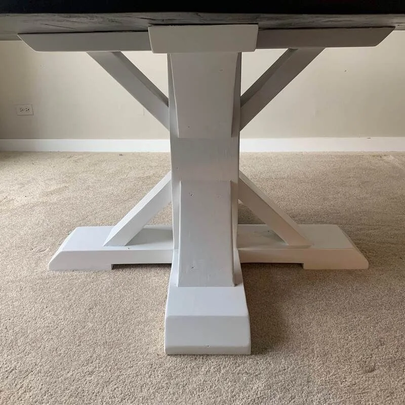 White wooden table base with an X-shaped support and wide flat bottom, photographed against a dark studio wall.