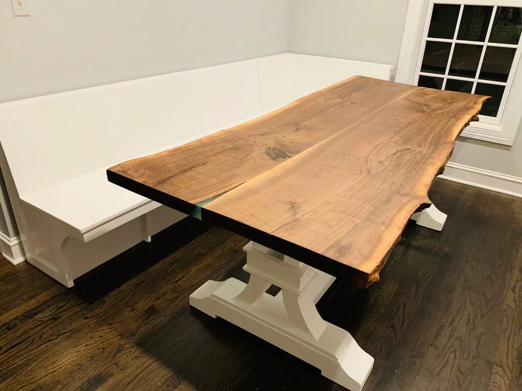 Live-edge wooden dining table with white pedestal legs and an L-shaped white bench, photographed against a dark studio wall.