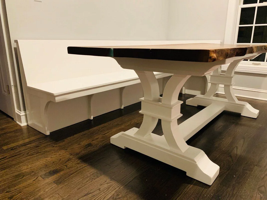 A thick natural wood table on white ornate pedestal legs, paired with a matching built-in white bench, photographed against a dark studio wall.