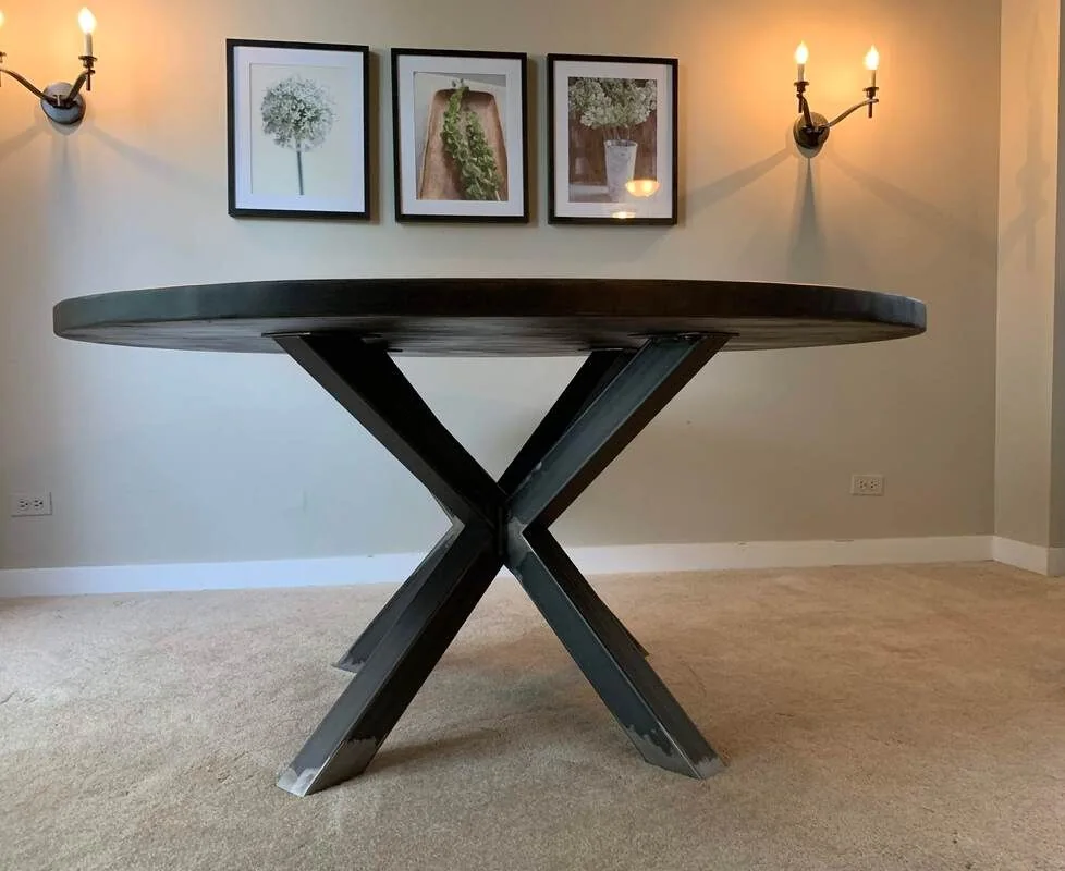 Round table with a dark top and X-shaped metal base on beige carpet, against a light gray wall with three floral pictures and two wall sconces.