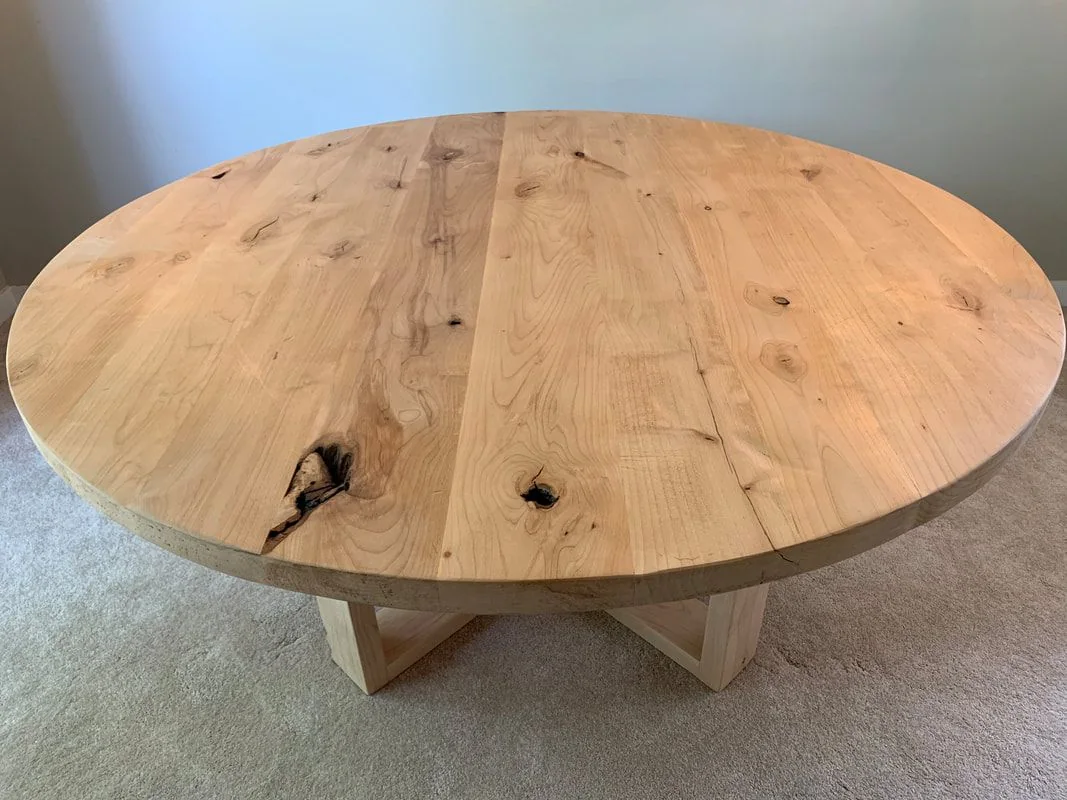 Round rustic wood table with intersecting leg base on carpeted floor.