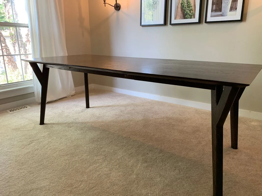 Dark wooden rectangular table with outward-angled legs in a carpeted room, positioned beneath three framed wall pictures and next to a window with white curtains.