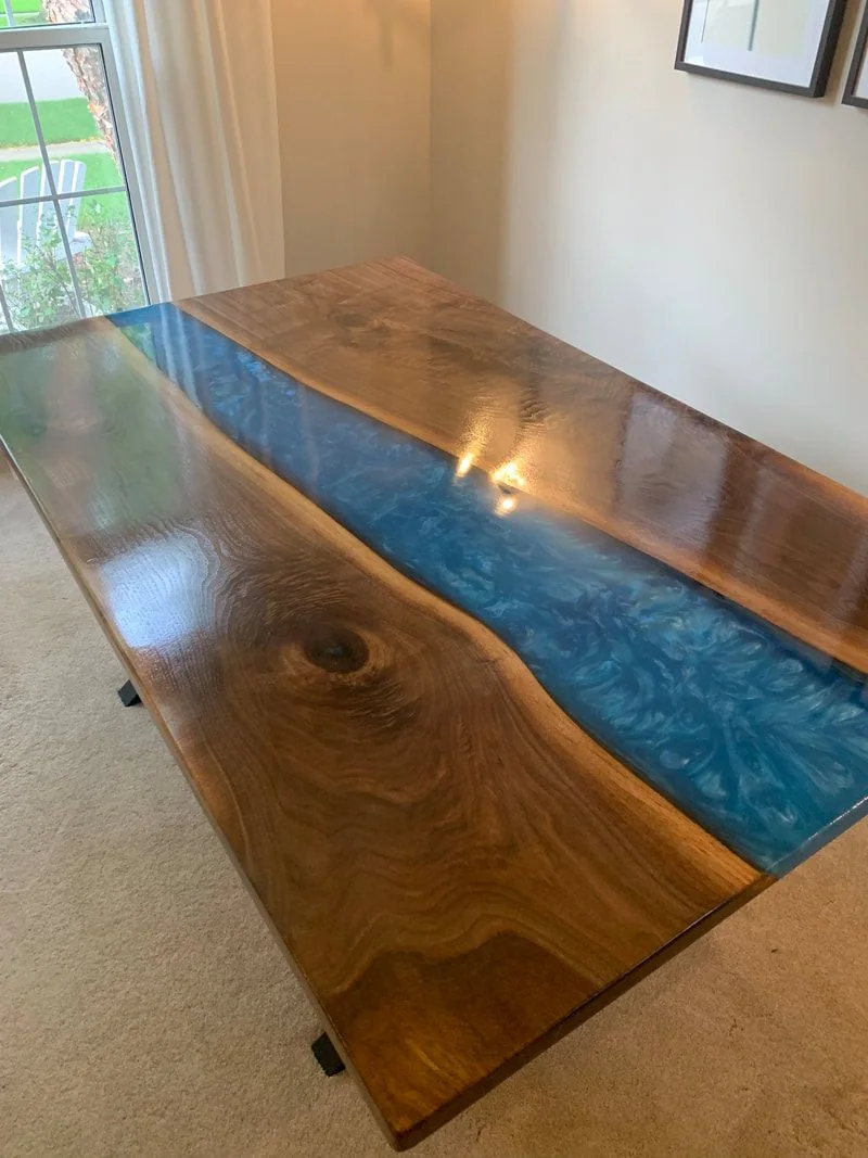 A wooden table with two polished slabs separated by a swirling blue resin river, placed on beige carpet near a window that highlights its glossy finish.