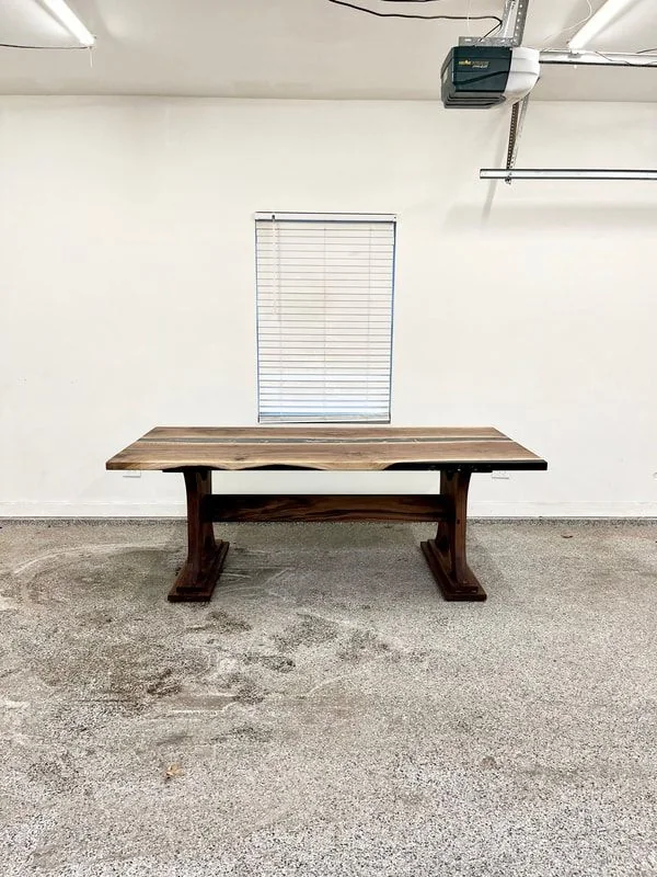 Wooden table with natural grain and sturdy legs connected by a horizontal beam, placed in a room with white walls, concrete floor, and a window with blinds.