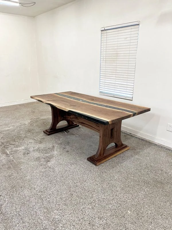 Wooden table with natural finish and two dark inlays, supported by curved legs, photographed against a dark studio wall.