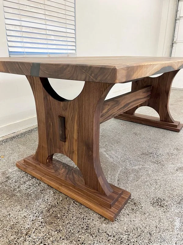 Dark wood table with thick rectangular top and arched legs featuring cut-out details, placed on a polished concrete floor in a room with white walls and blinds.