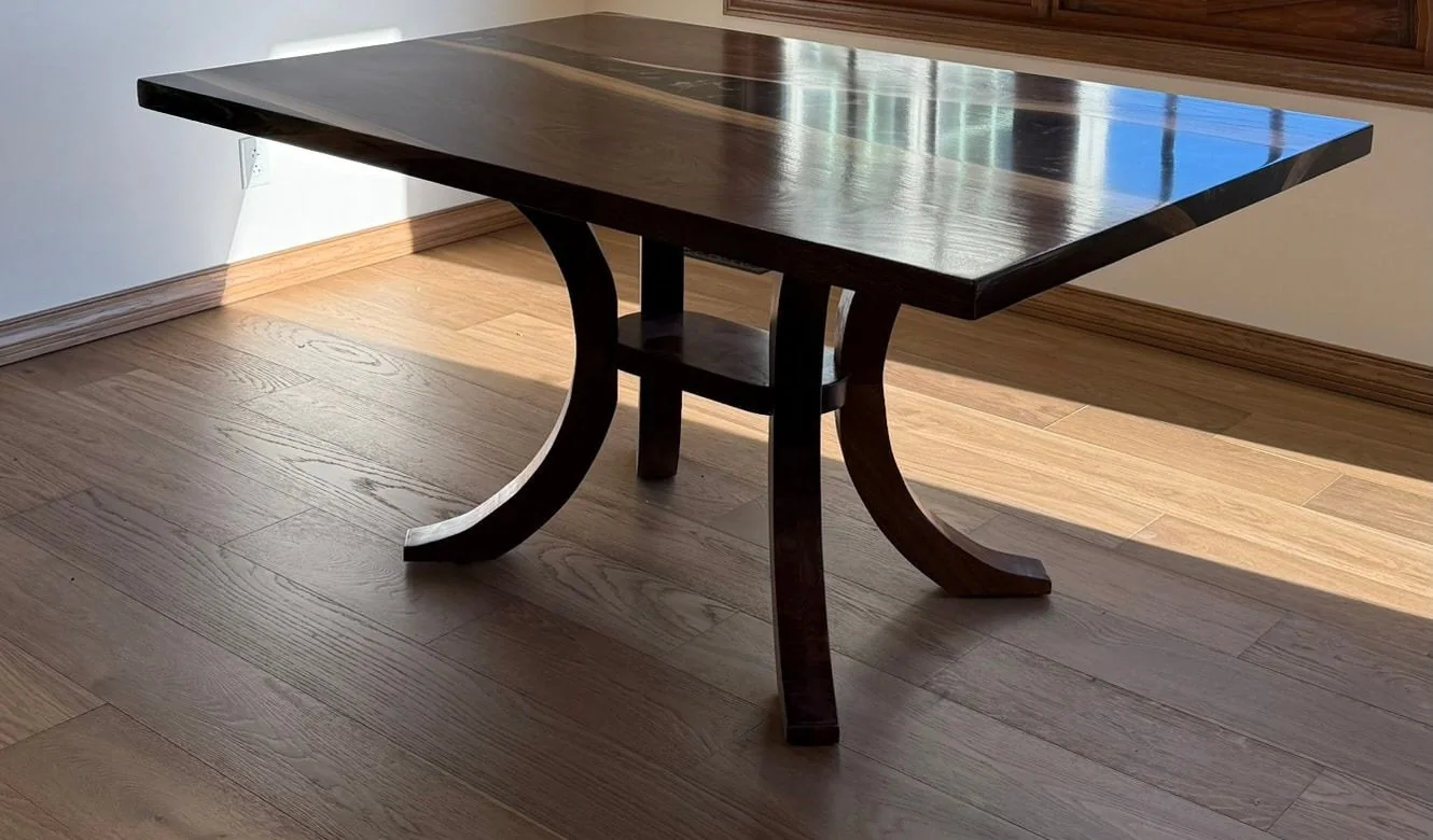A dark walnut dining table with a narrow resin channel, standing on a pedestal of four curved walnut legs joined by a small central shelf, on a pale oak floor in afternoon light.