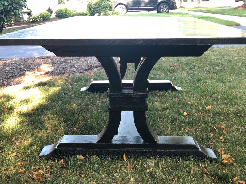 Dark wooden table with decorative curved base, placed on a grassy lawn with a driveway and greenery in the background.