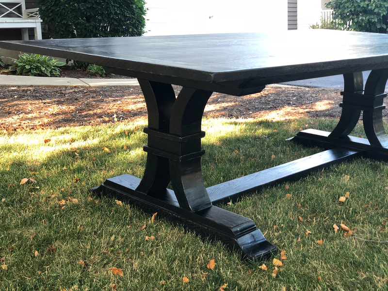 Black wooden table with rectangular top and curved legs connected by a horizontal beam, placed on a grassy lawn with greenery in the background.