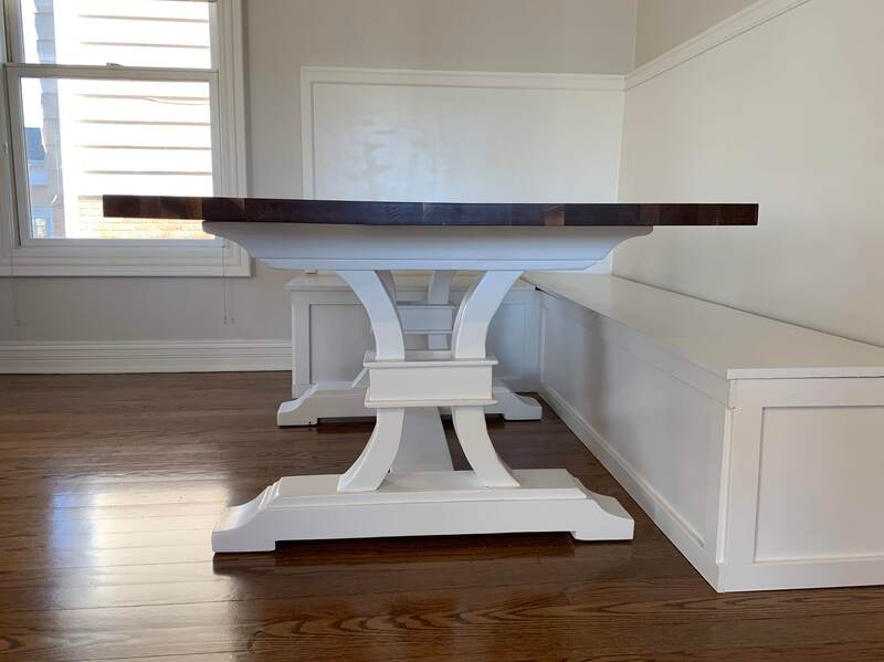 Dark wood dining table with white ornate pedestal legs, paired with built-in white bench seating in a cozy dining nook with hardwood flooring and natural light