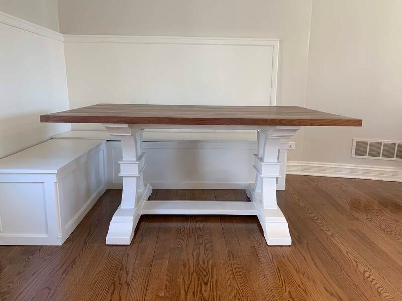 Rectangular wooden table with white pedestal legs, placed on hardwood flooring next to built-in white bench seating in a modern dining area.