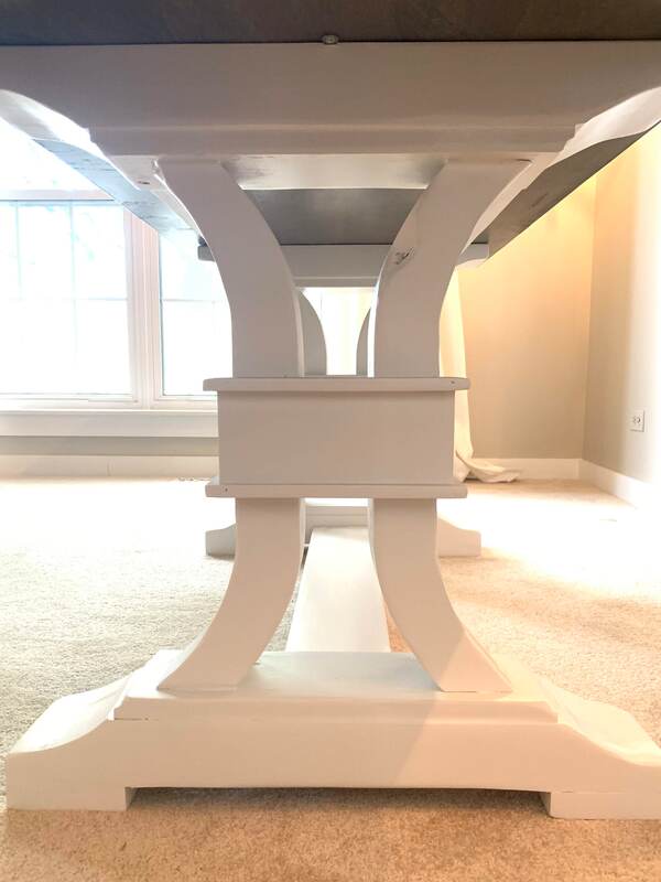 Underside of a table with white curved supports and a horizontal connector, placed on a carpeted floor with natural light from a nearby window.