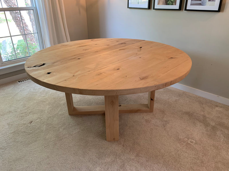 Round rustic wood table with intersecting square base on beige carpet, framed wall art and window with white curtains in background.