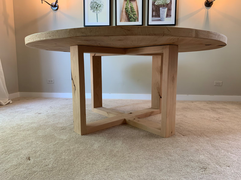 Round wood table with thick top and Open Looped base on carpeted floor, framed floral art and wall lights in background.